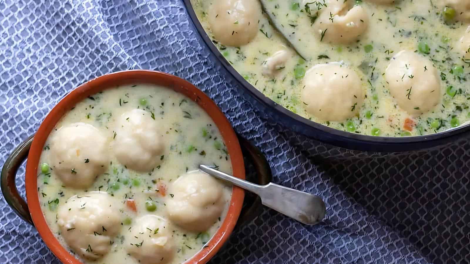Bowl and pot filled with creamy soup featuring round dumplings, peas, carrots, and herbs on a textured surface.