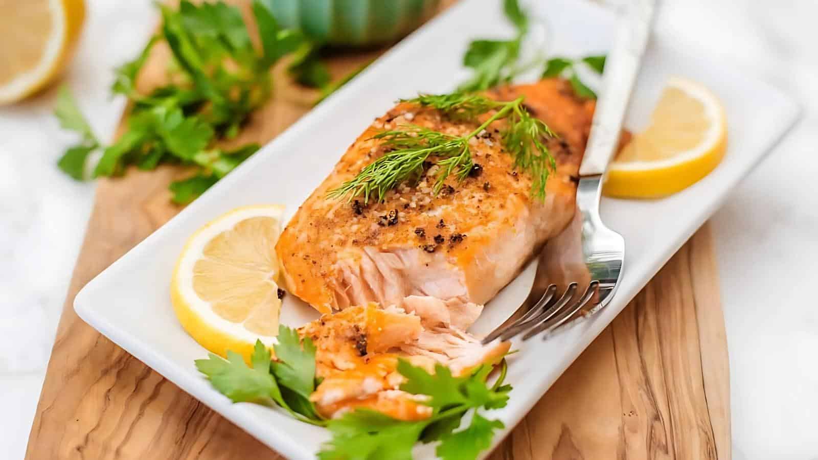 Grilled salmon fillet garnished with dill and black pepper on a white plate, accompanied by lemon slices and parsley, with a fork placed beside it.
