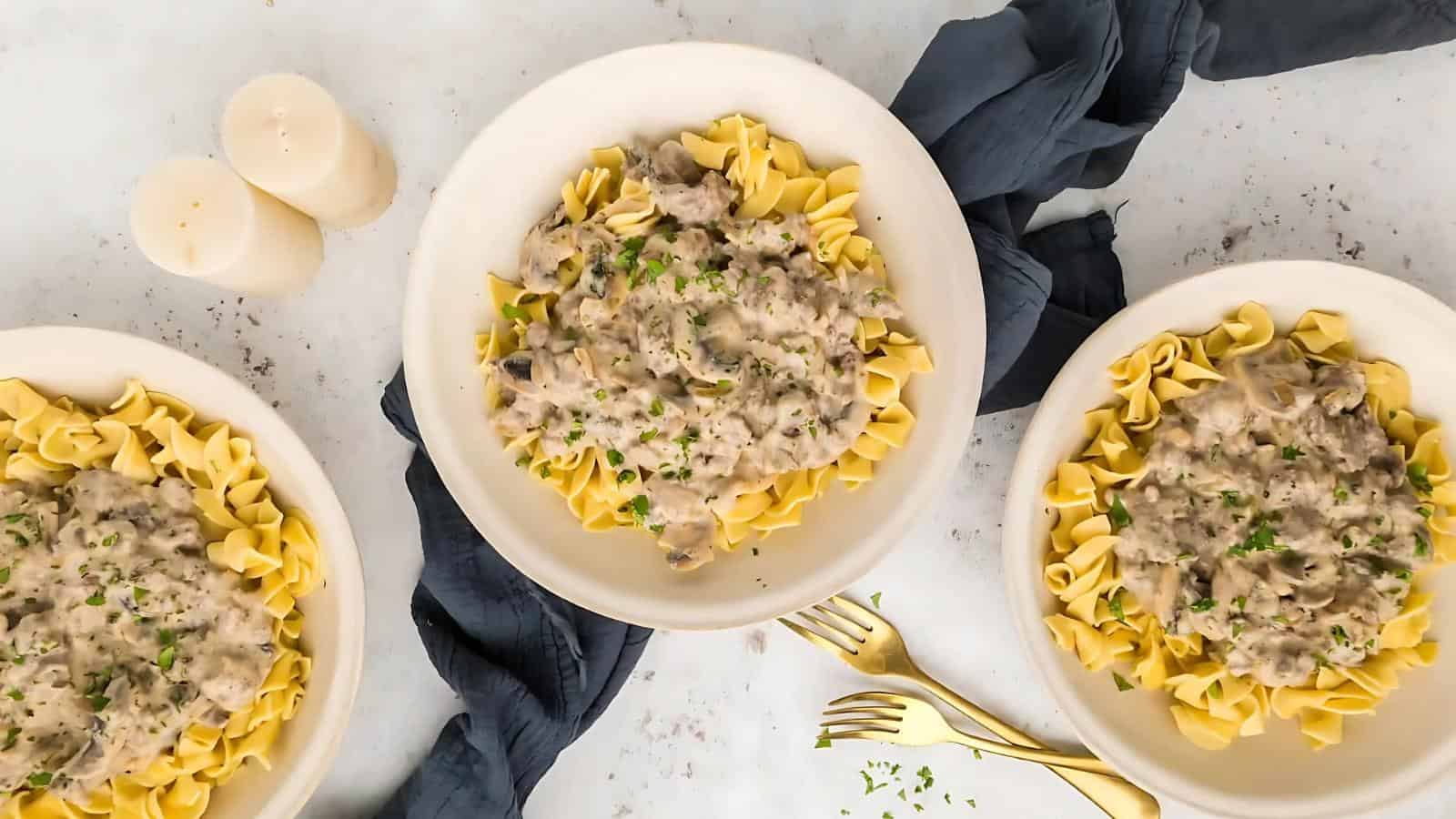 Three bowls of pasta topped with creamy mushroom sauce are arranged on a table with a dark cloth, candles, and gold forks.