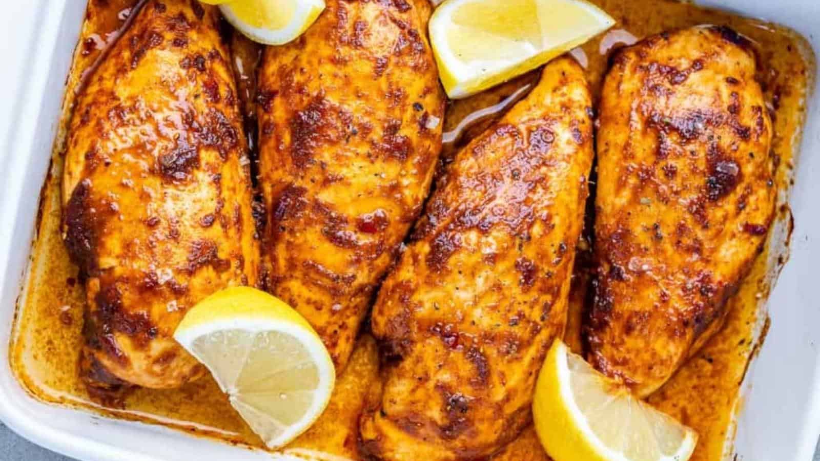 Four cooked chicken breasts seasoned with spices and garnished with lemon wedges in a white baking dish.