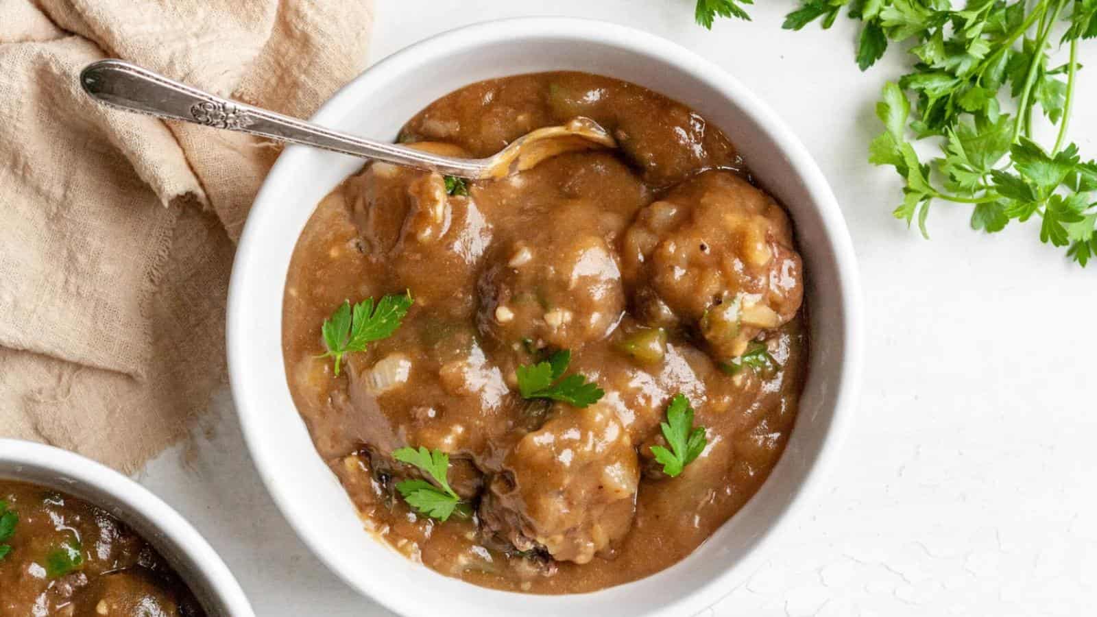 A bowl of brown stew with dumplings and parsley garnish, placed on a light surface with a silver spoon.