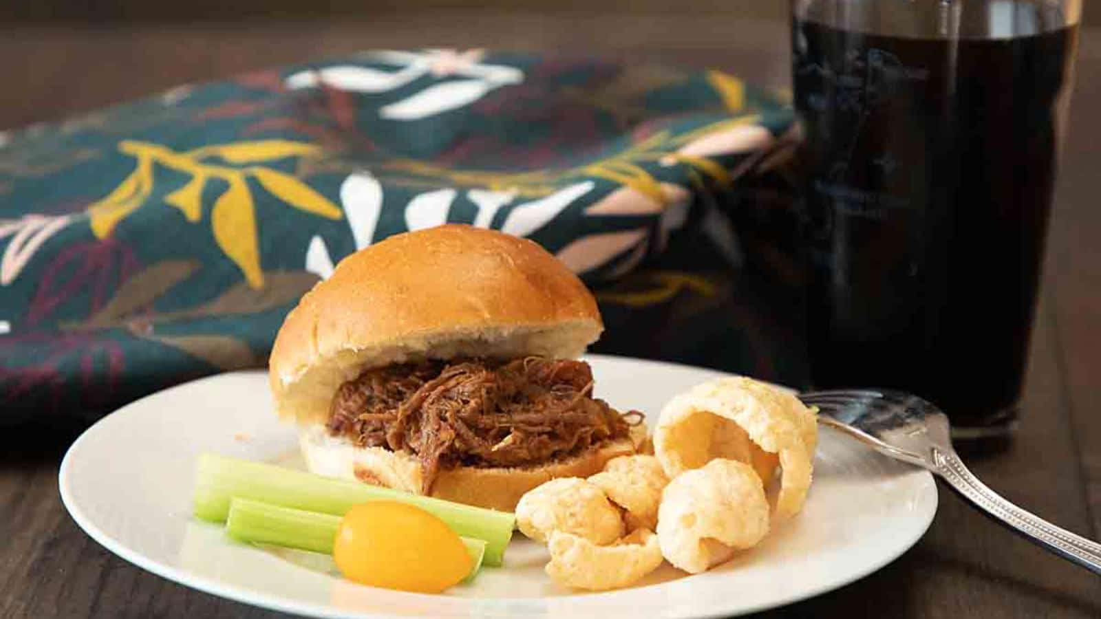 Pulled pork sandwich on a plate with celery sticks, cheese puff snack, and a cherry tomato. A colorful napkin and a glass of dark beverage in the background.