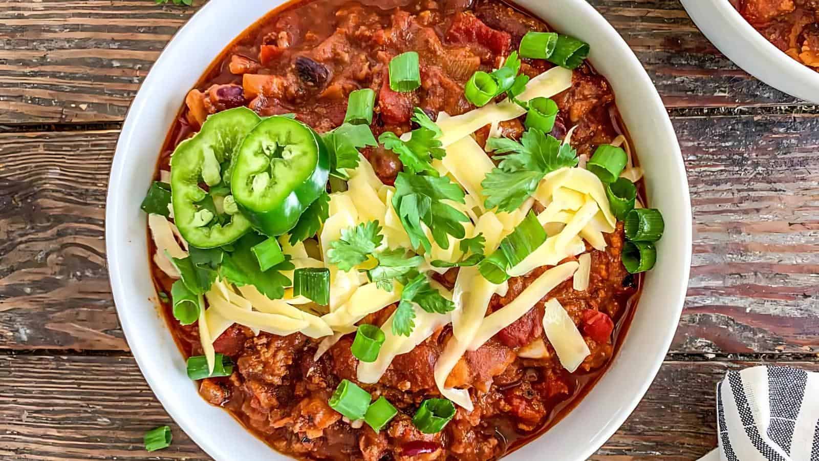 A bowl of chili topped with shredded cheese, sliced jalapeños, chopped green onions, and cilantro on a wooden table.