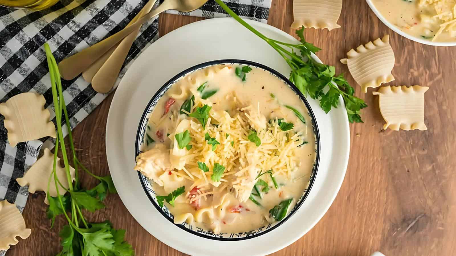 A bowl of creamy soup with shredded cheese, chicken, and herbs on a plate. Decorative pasta pieces and fresh parsley are scattered on a wooden table with a checkered cloth and cutlery.