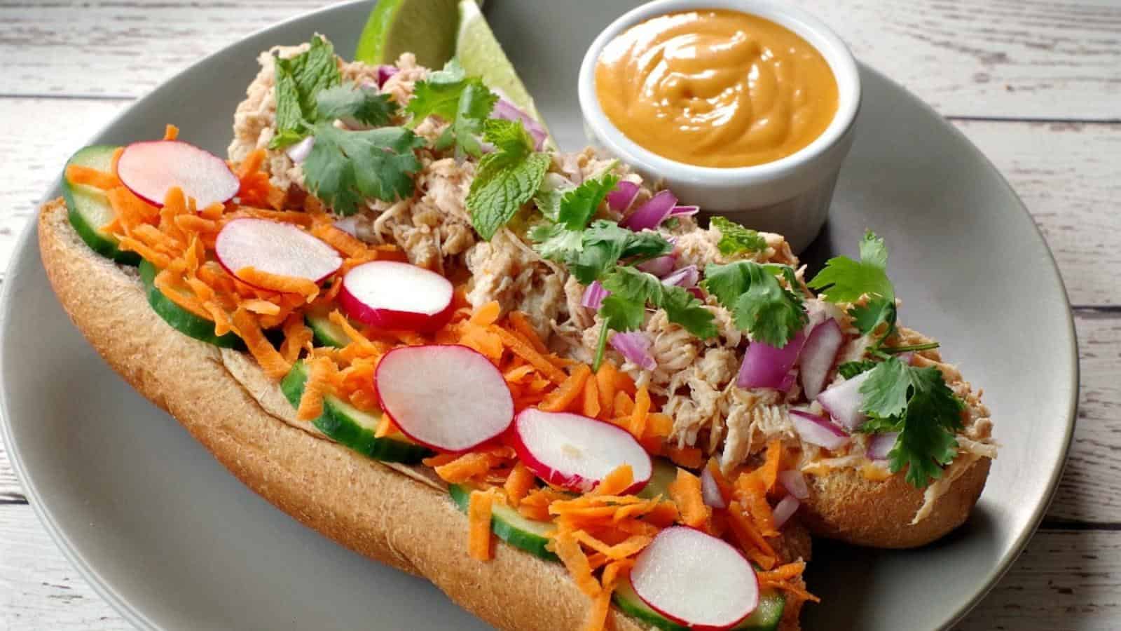 A sandwich with shredded chicken, cucumber, radish, carrot, cilantro, and onions on a plate, accompanied by a small bowl of dipping sauce.