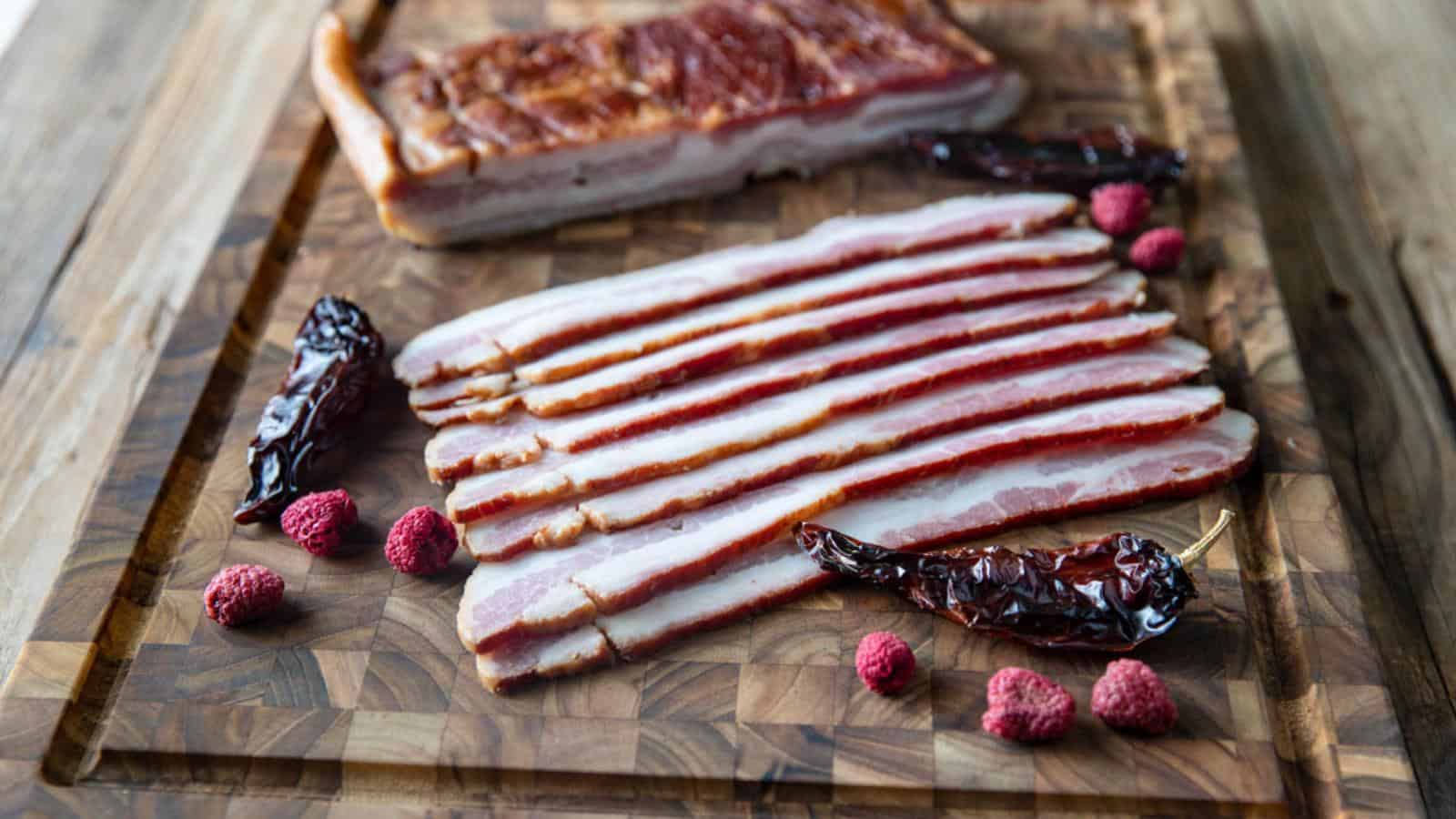 Slices of bacon on a wooden board with dried chilies and raspberries.