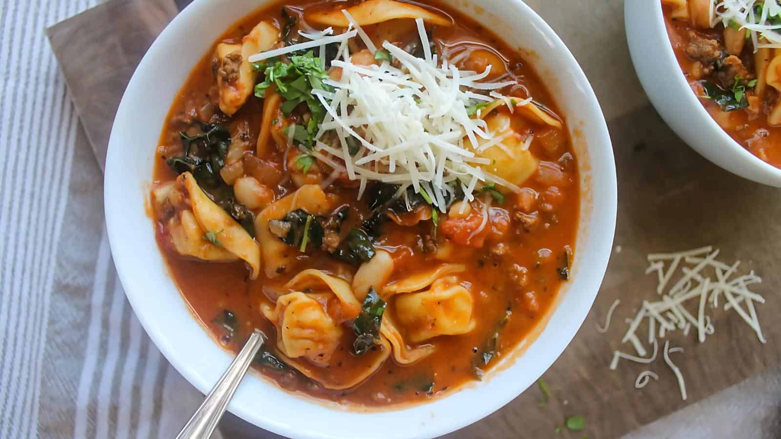 Bowl of tomato-based tortellini soup topped with grated cheese and herbs, with a spoon on the side and cheese scattered on a wooden surface.