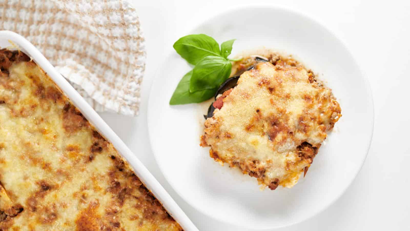Slice of lasagna with basil on a white plate next to a casserole dish filled with lasagna.