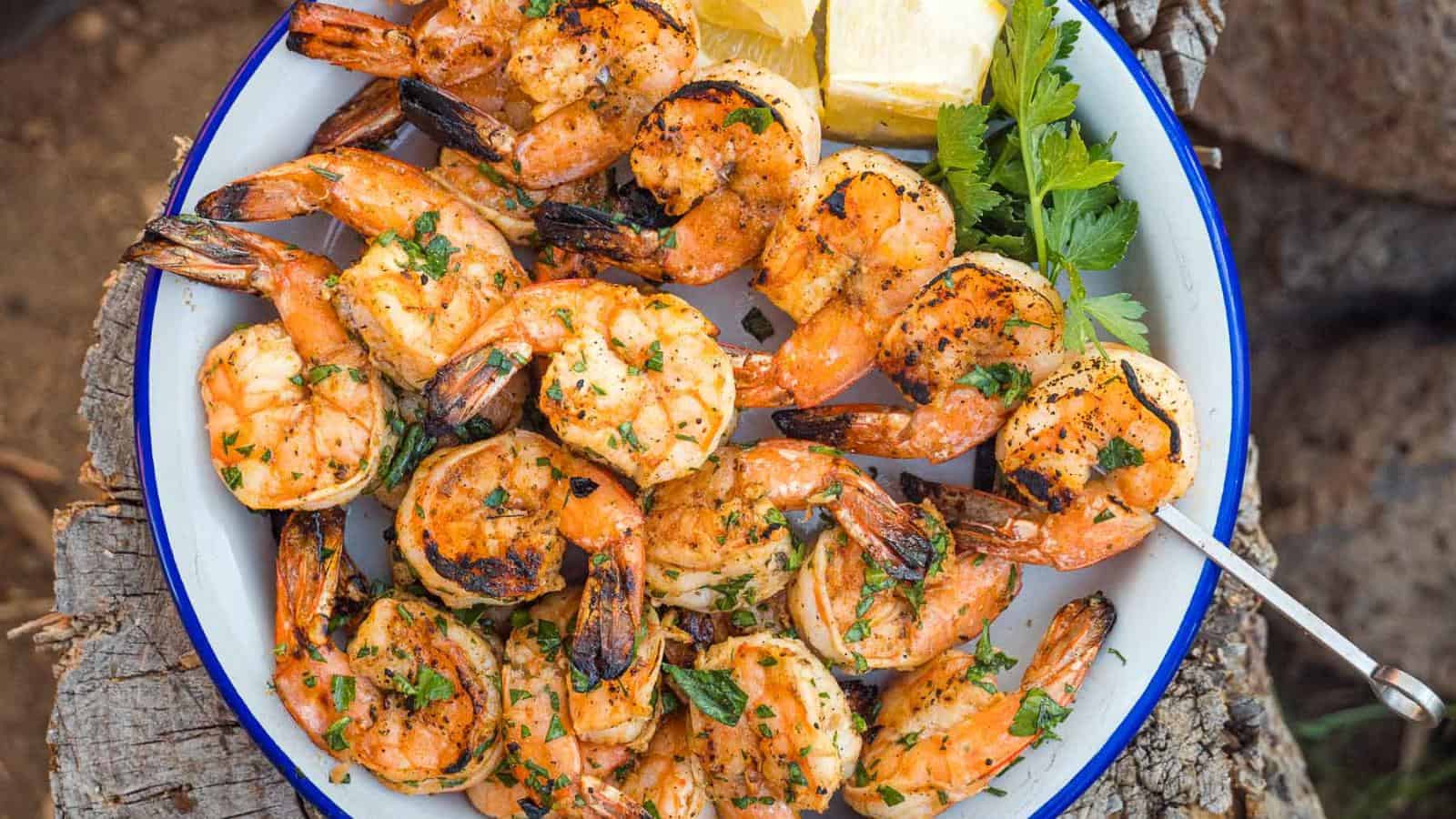 A plate of grilled shrimp garnished with herbs, lemon wedges, and a skewer, served on a rustic wooden surface.