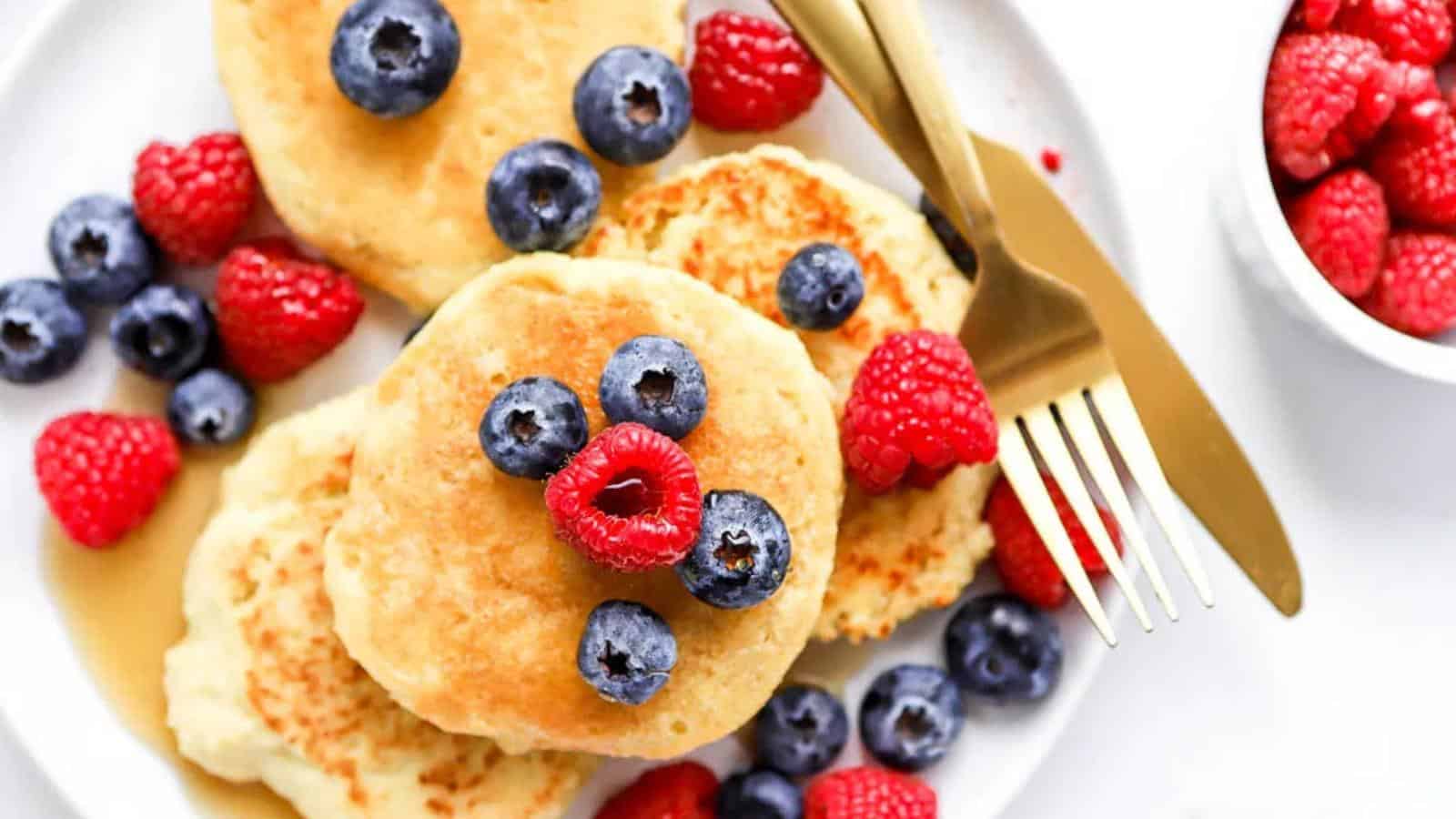 Plate of pancakes topped with blueberries and raspberries, with a fork and knife beside. Additional raspberries in a small bowl.