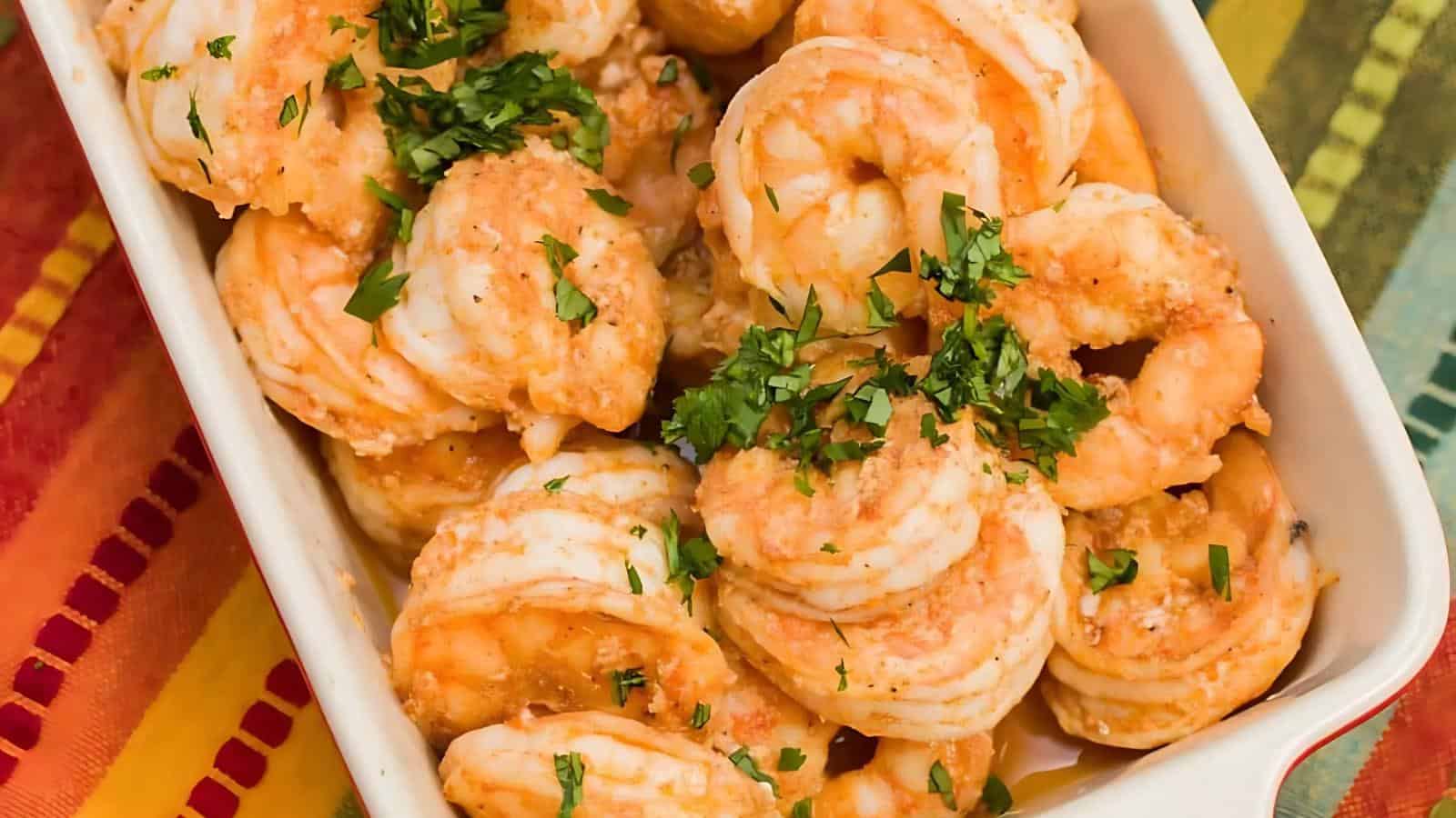 A rectangular dish filled with seasoned cooked shrimp garnished with chopped parsley, set on a colorful striped tablecloth.