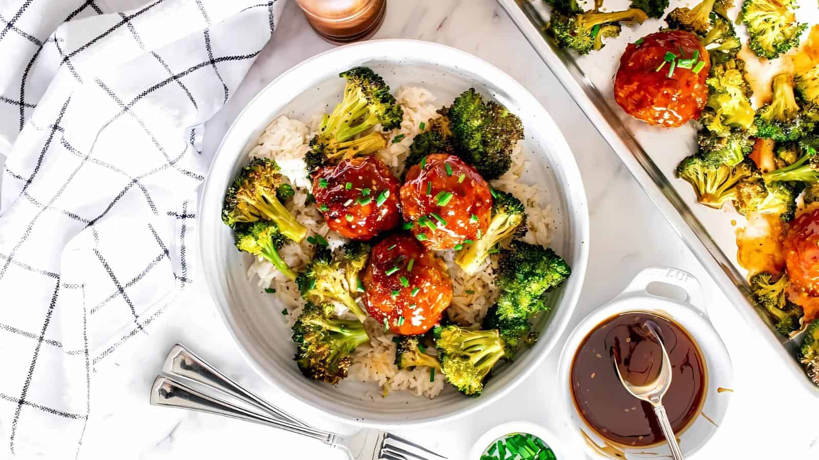 A bowl of rice topped with broccoli and glazed meatballs, garnished with green onions, is set on a table with a checkered napkin and utensils. There's a small dish of sauce nearby.