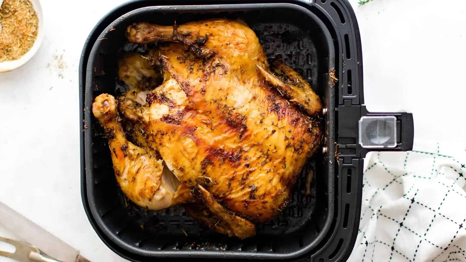 Whole roasted chicken inside an air fryer basket, seasoned with herbs and spices, on a white surface with a green cloth.