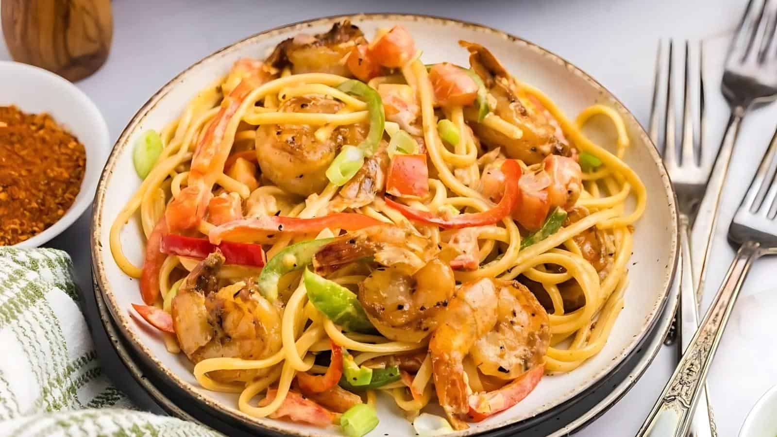 A plate of shrimp and pasta mixed with sliced red and green bell peppers, garnished with green onions. Two forks and a napkin are placed beside the plate.
