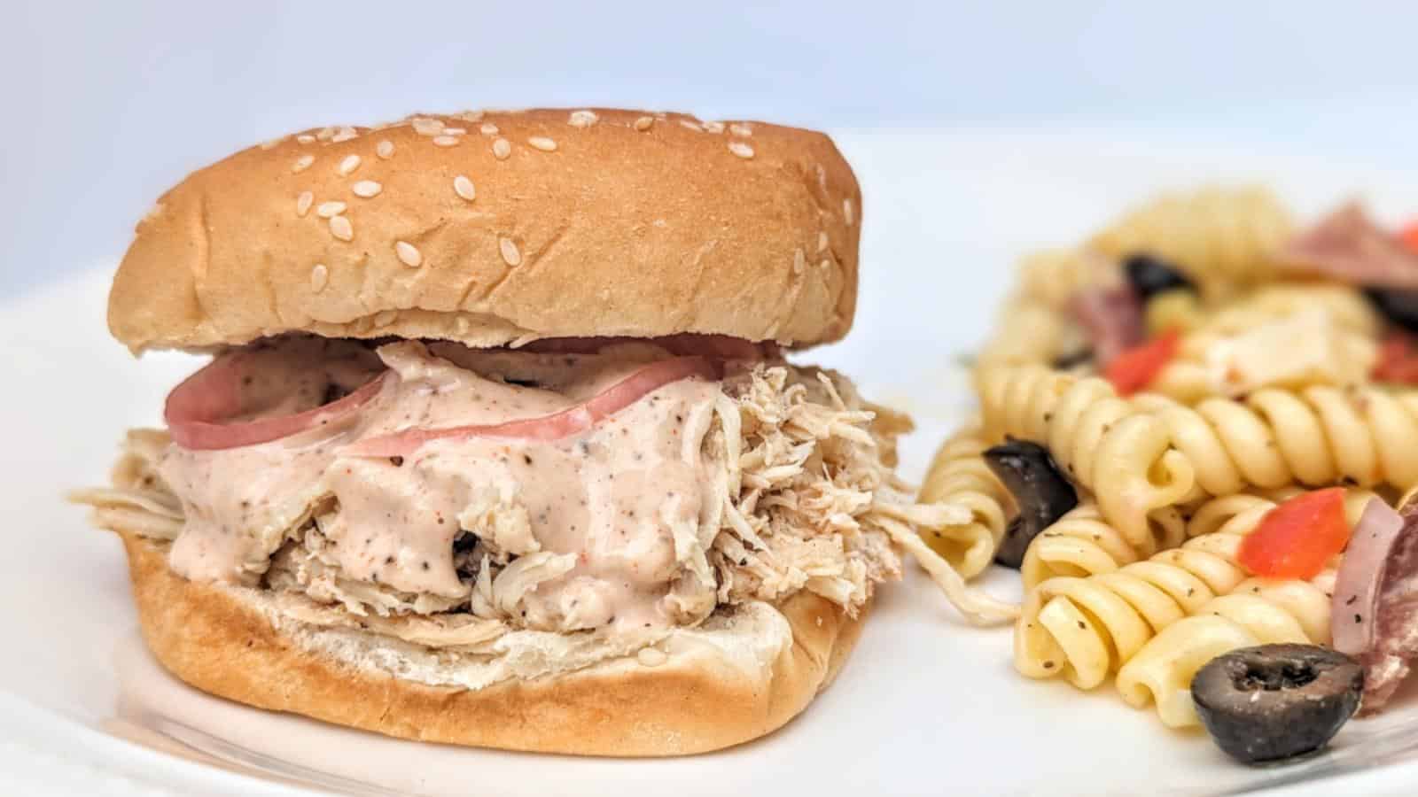 Shredded chicken sandwich with creamy sauce and pickled onions on a sesame bun, served with a side of rotini pasta salad with olives, tomatoes, and peppers.
