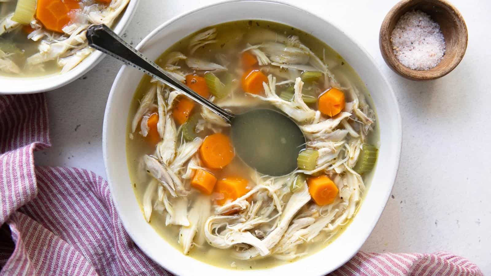 A bowl of chicken soup with carrots and celery, alongside a spoon. A small bowl of salt is nearby, with a red-striped cloth to the side.