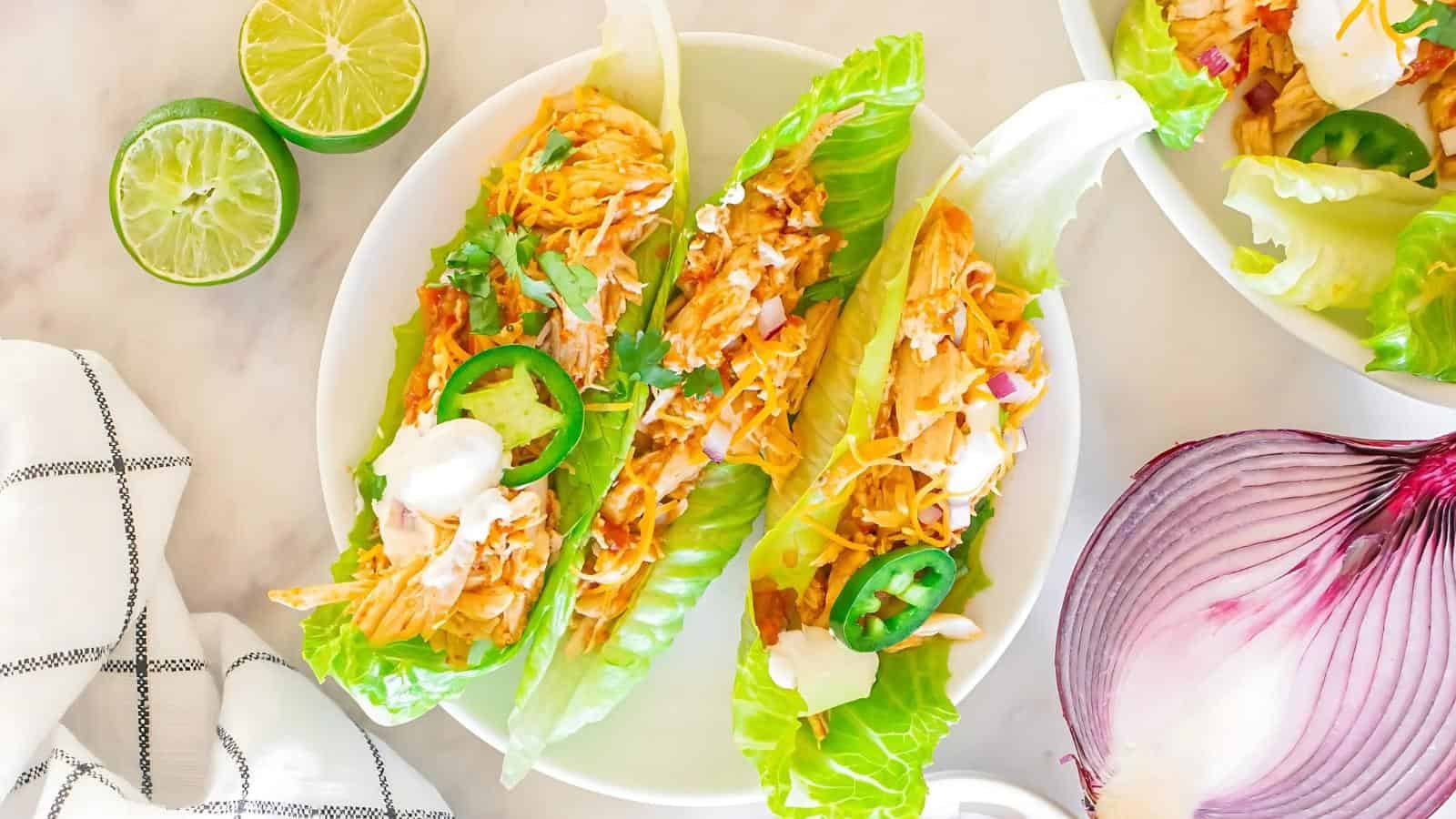 Lettuce wraps filled with shredded chicken, sliced jalapeños, cheese, and sour cream on a plate. Lime halves and a red onion are nearby.