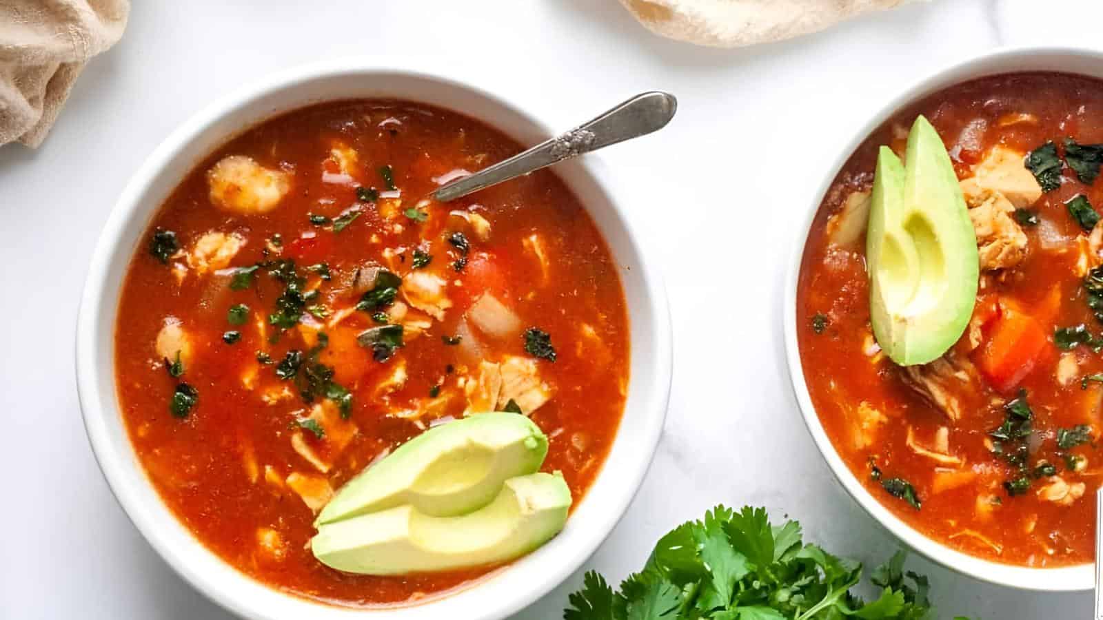 Two bowls of tomato-based soup with chicken chunks, avocado slices, and cilantro garnish. A spoon rests in one bowl. Fresh cilantro is scattered nearby.