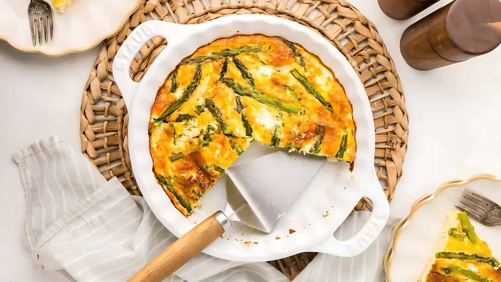 Quiche with asparagus and spinach in a white baking dish, with one slice removed, on a woven placemat. Two forks on striped napkins and salt and pepper shakers are nearby.