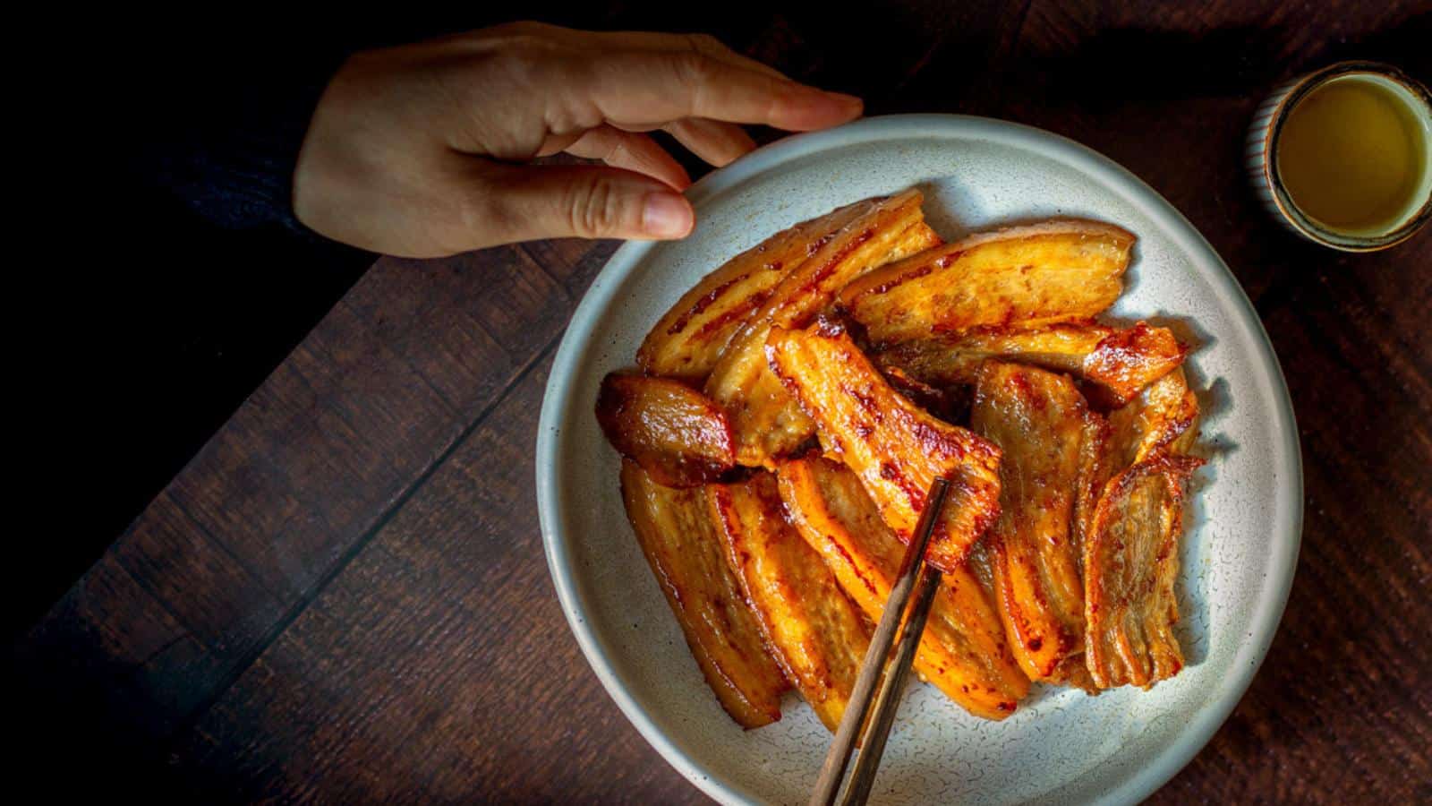 A hand reaches for a plate of grilled pork belly slices on a dark wooden table, with chopsticks resting on the plate and a small cup of yellow sauce nearby.