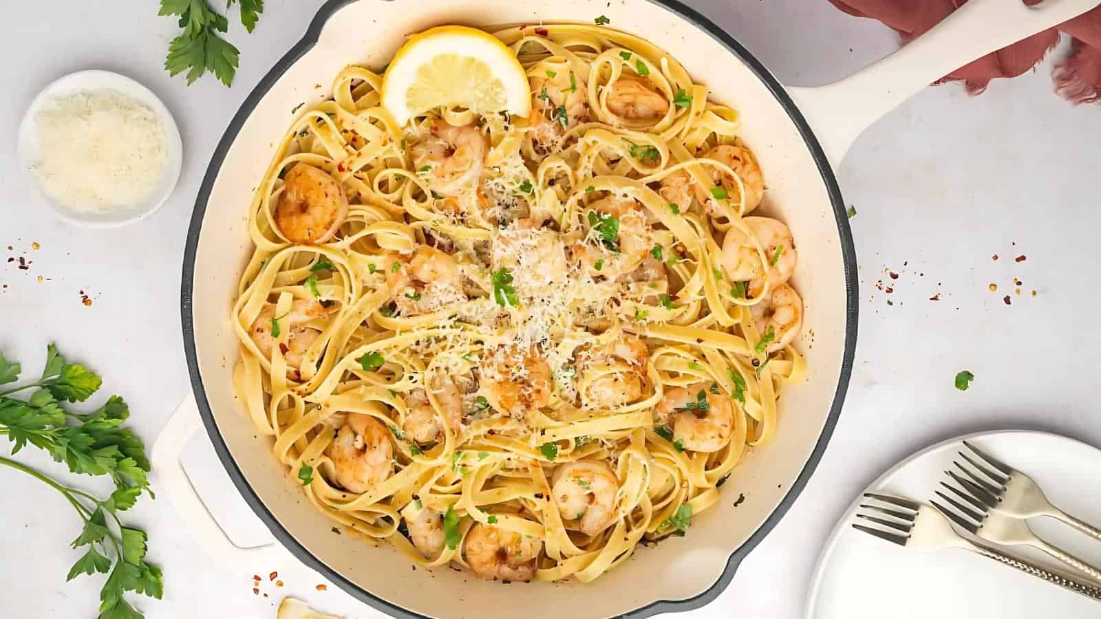 A white skillet filled with shrimp linguine topped with grated cheese and parsley, garnished with a lemon slice. Nearby, there are plates, forks, parsley, red pepper flakes, and grated cheese.