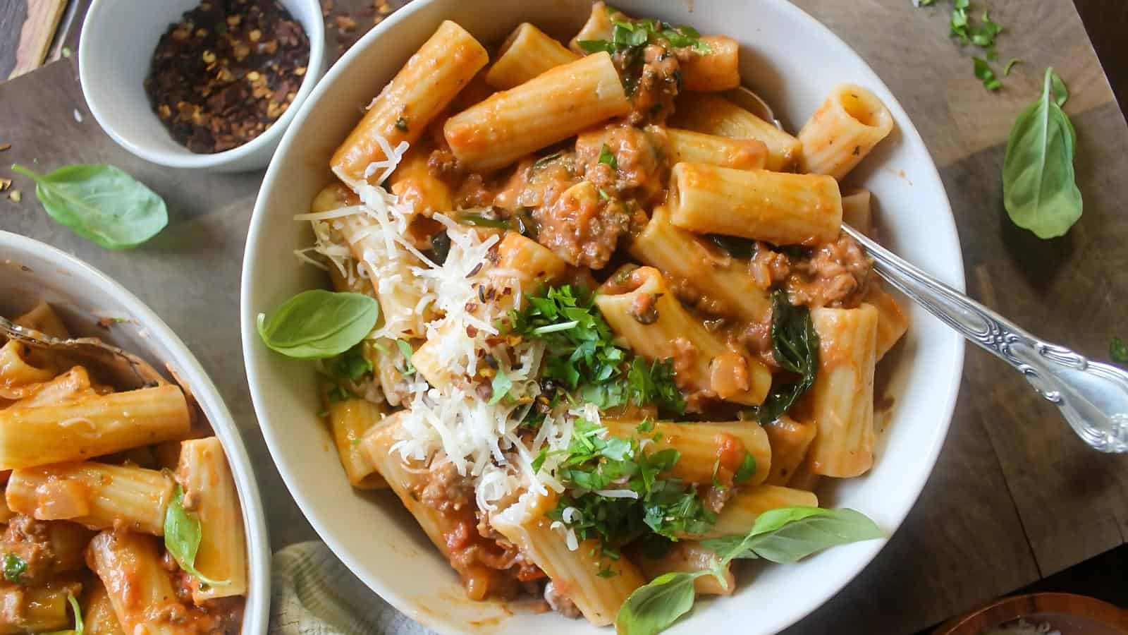 A bowl of rigatoni pasta with meat sauce, topped with grated cheese and fresh basil leaves, set on a wooden surface. A bowl of red pepper flakes is in the background.