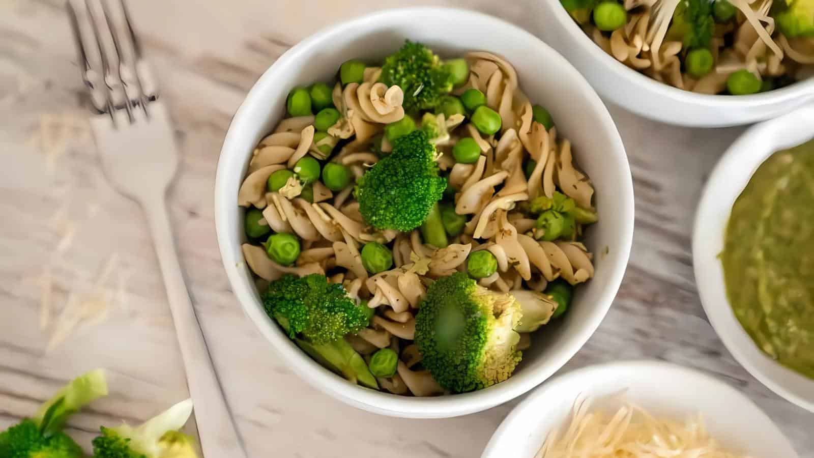 Bowl of pasta with broccoli and peas next to a dish of sauce and a plate of grated cheese on a table.