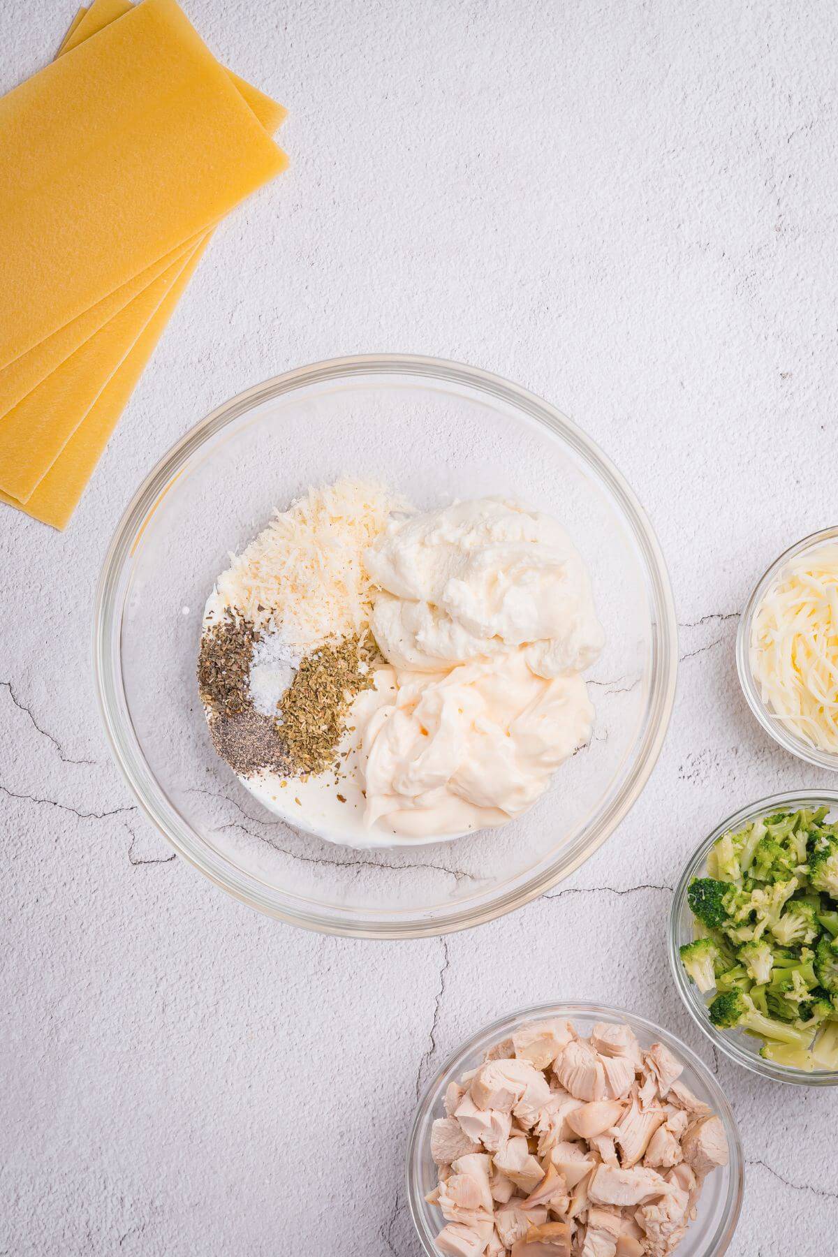 A bowl with cheese, herbs, and cream mixtures surrounded by bowls of chopped chicken, zucchini, shredded cheese, and lasagna sheets on a white surface.
