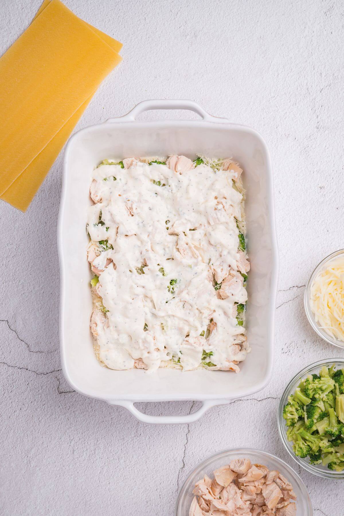 White baking dish with lasagna being assembled, featuring white sauce, shredded chicken, and broccoli. Nearby are lasagna sheets and bowls with more ingredients.
