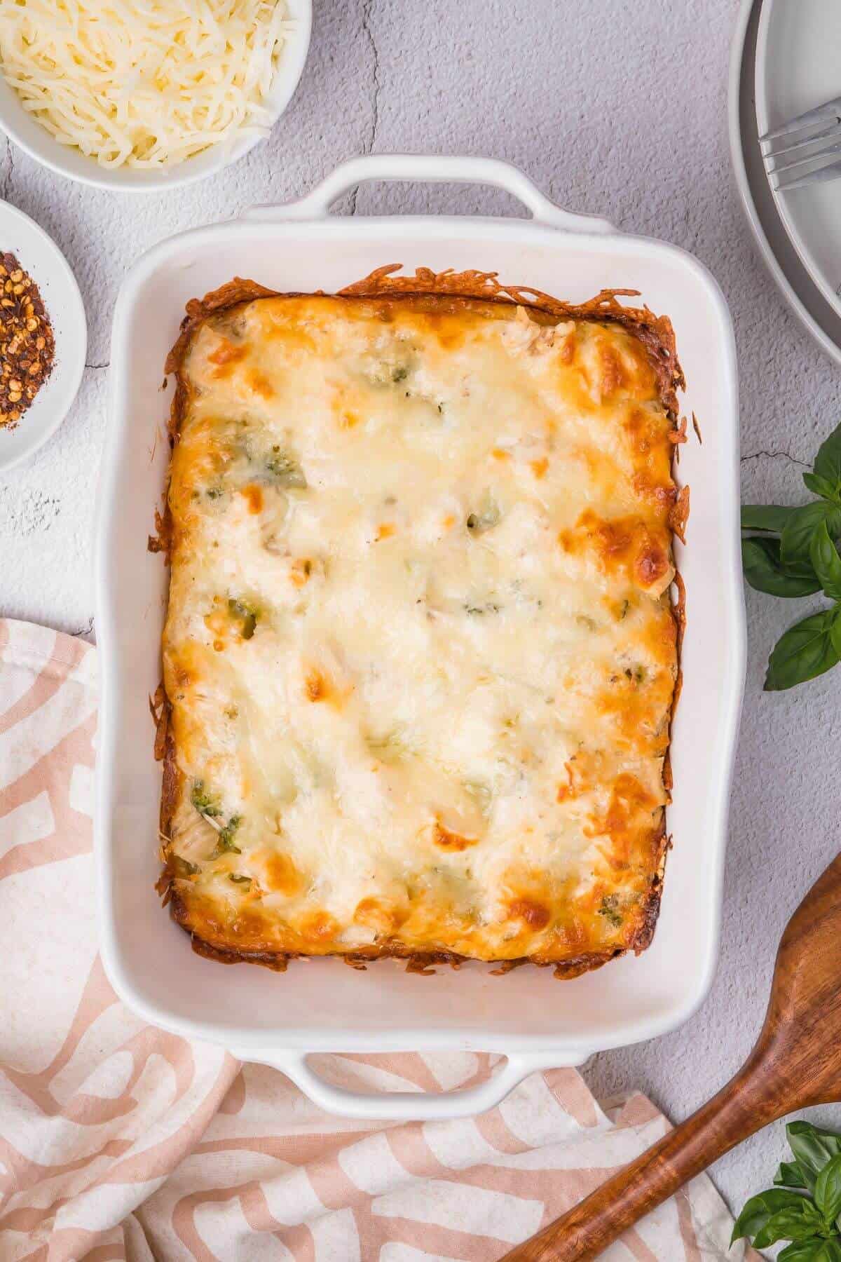 A baked lasagna in a rectangular dish on a counter, surrounded by a wooden spoon, a bowl of shredded cheese, chili flakes, and a beige patterned cloth.