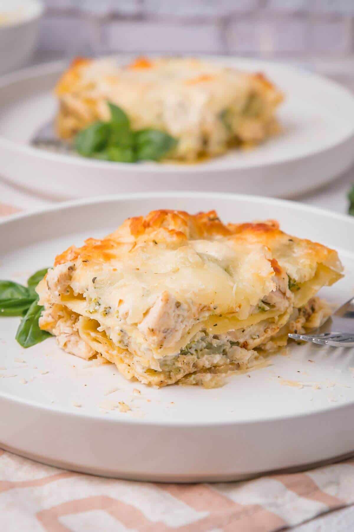 A slice of chicken and spinach lasagna on a white plate with a fork and basil garnish.