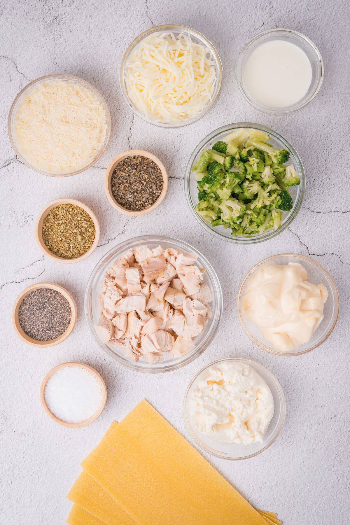 Ingredients on a table: shredded cheese, cream, chopped broccoli, herbs, spices, diced chicken, mayonnaise, ricotta, and lasagna sheets.