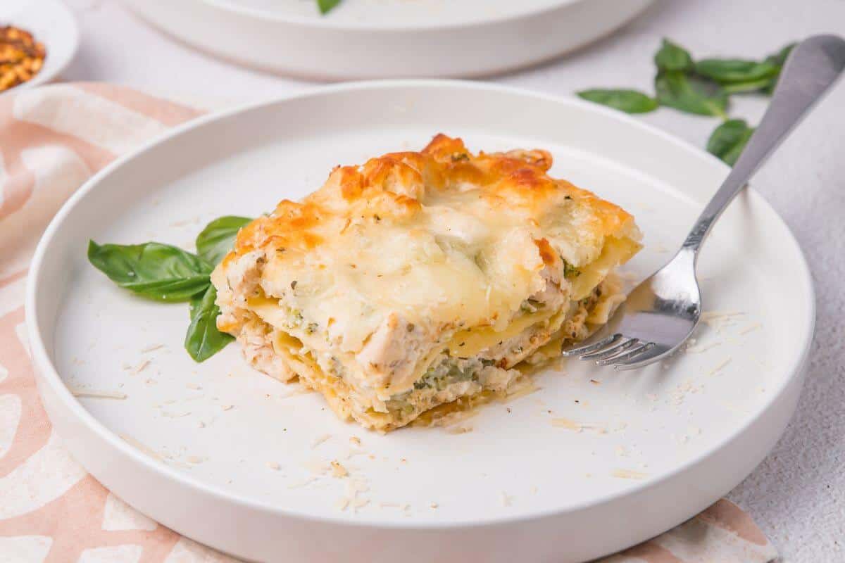 A slice of cheesy lasagna with visible layers of pasta and filling on a white plate, garnished with basil leaves. A fork rests beside the lasagna.