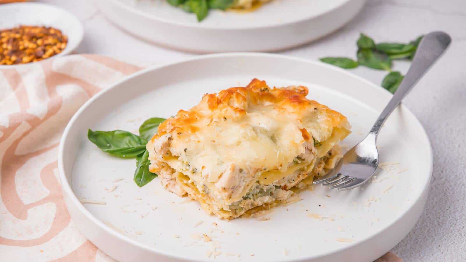 A slice of lasagna on a white plate, garnished with basil leaves, and a fork beside it.