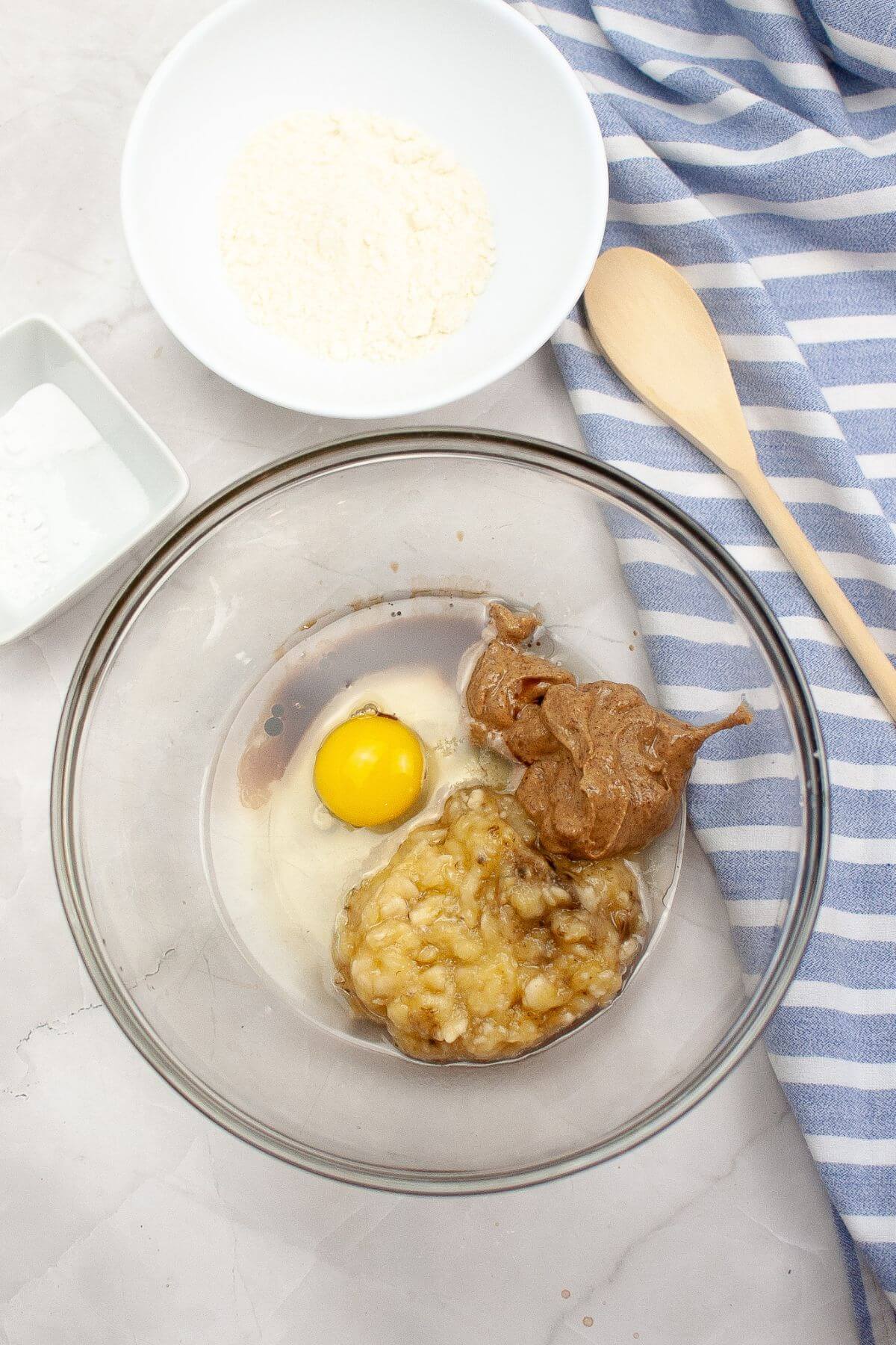 Ingredients in a glass bowl: mashed banana, egg, and peanut butter. Nearby are bowls with flour, baking soda, and a wooden spoon on a striped cloth.