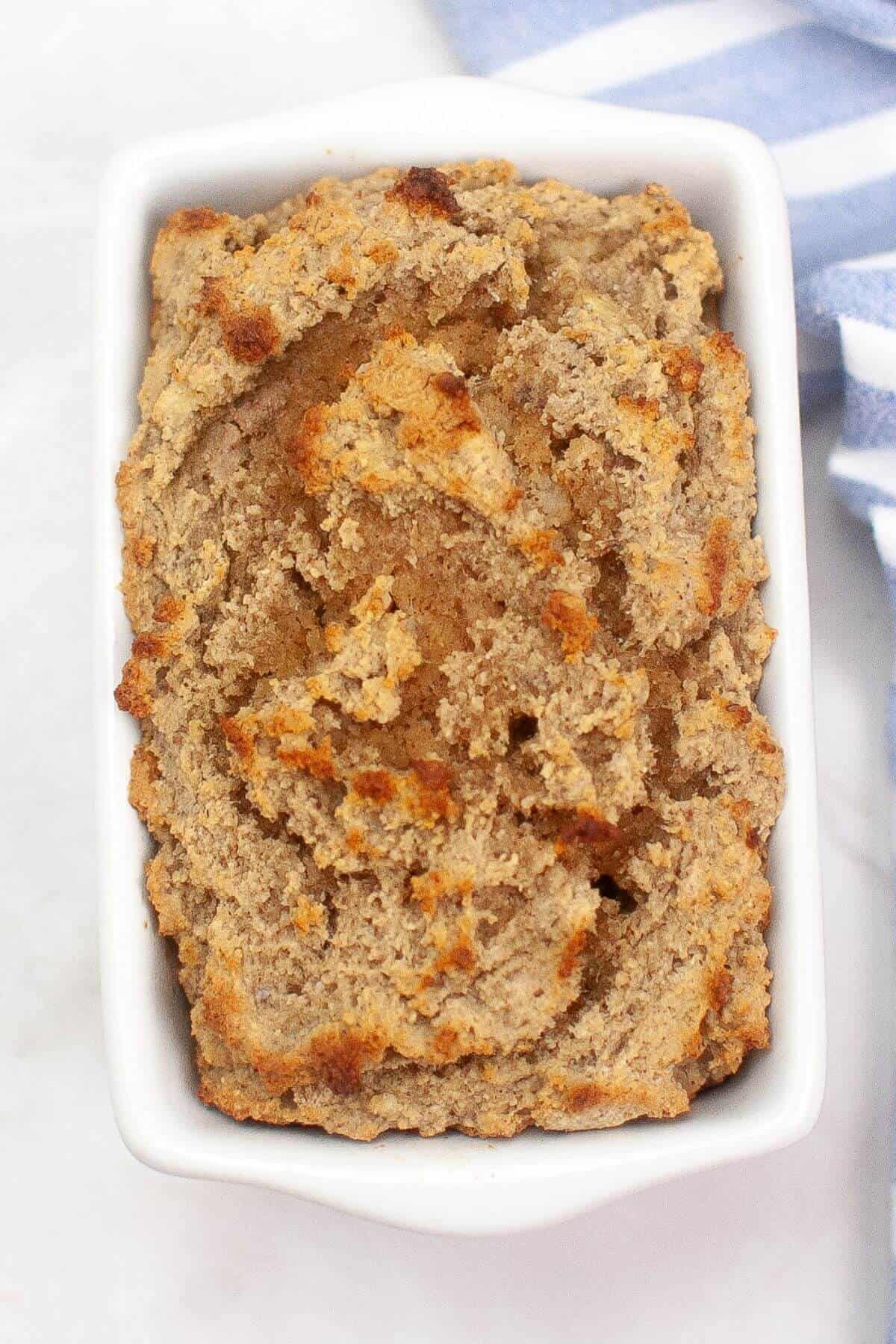 A rectangular white dish filled with freshly baked banana bread, featuring a golden-brown, textured crust. A folded striped cloth is partially visible beside it.