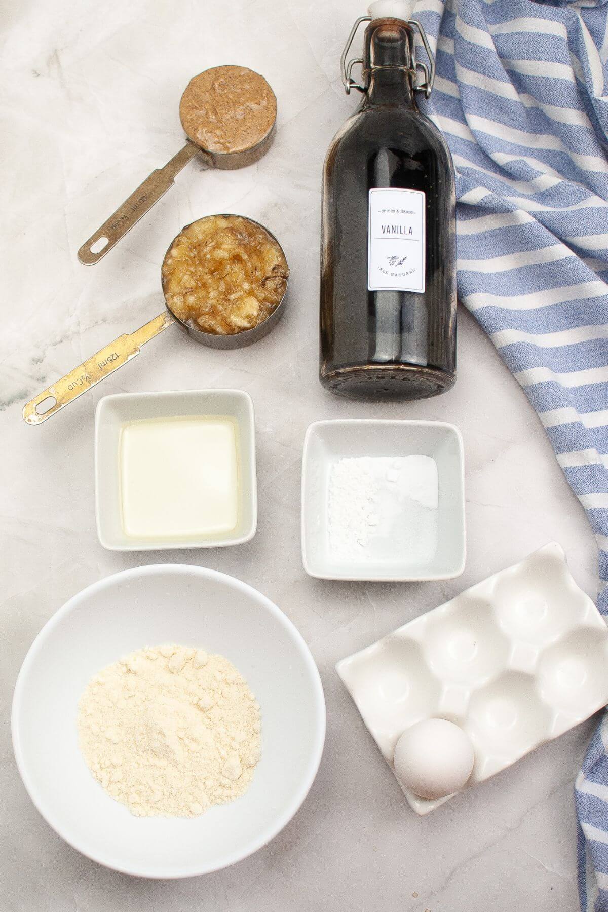 Ingredients on a countertop: vanilla extract bottle, mashed bananas, peanut butter, flour, egg, oil, baking powder, and a striped cloth.