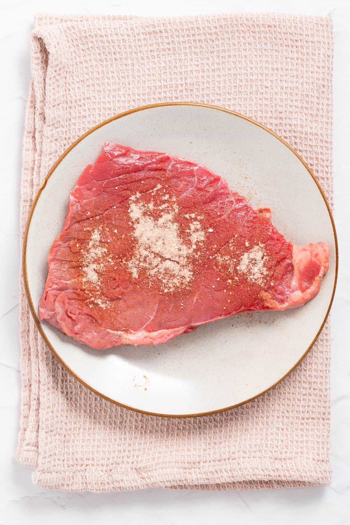 Raw steak with seasoning on a round plate, placed on a pink textured cloth.