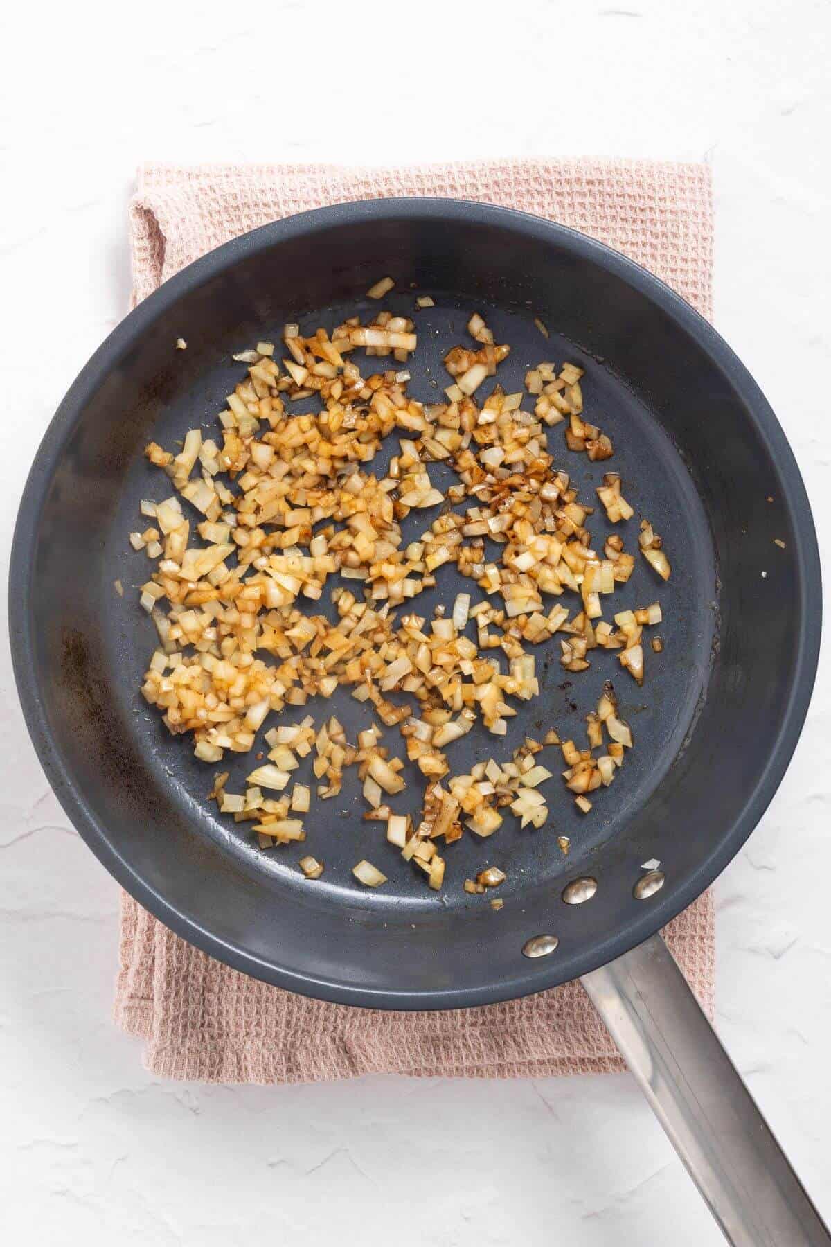Chopped onions are being sautéed until golden brown in a frying pan placed on a textured towel.