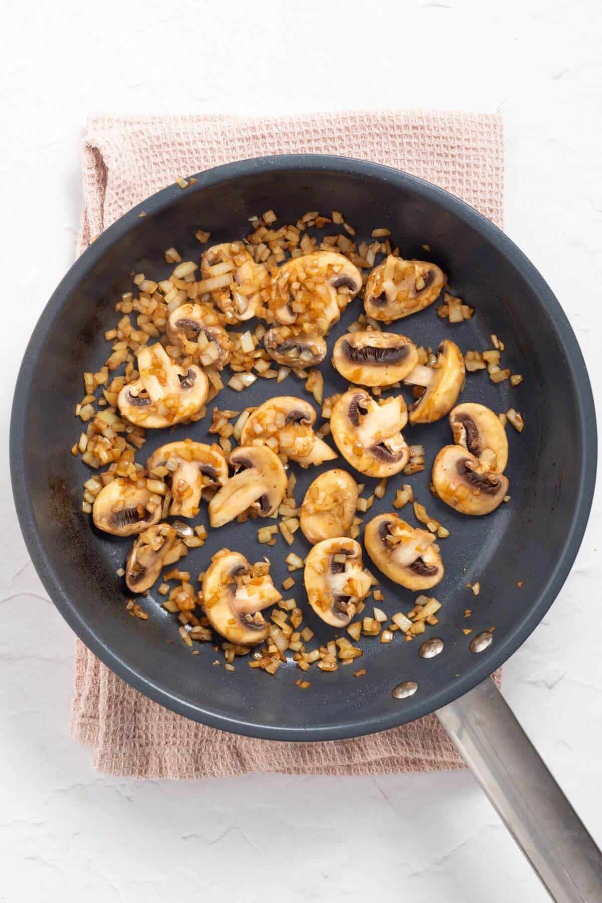 Sliced mushrooms and chopped onions sautéing in a black frying pan on a light pink cloth.