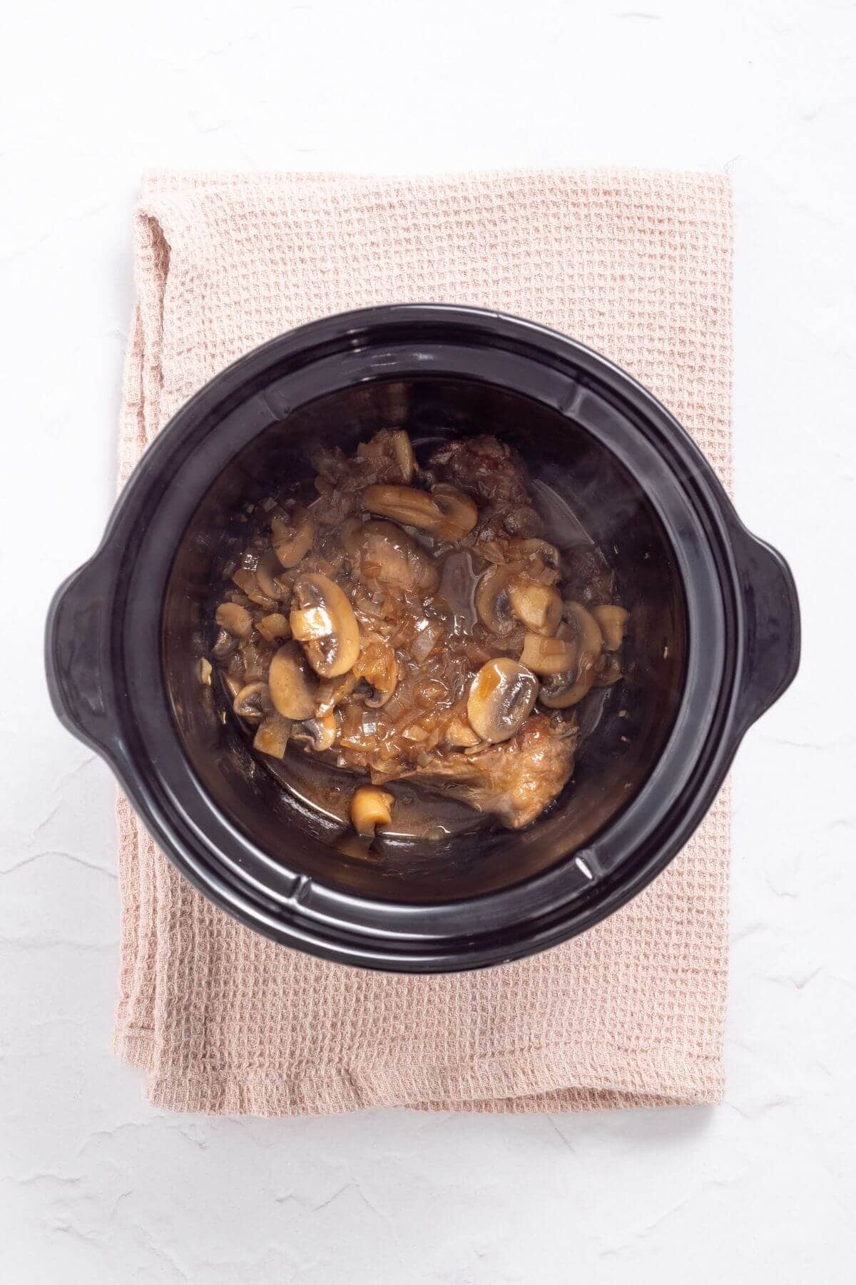A slow cooker with cooked chicken and mushrooms, placed on a folded beige towel.