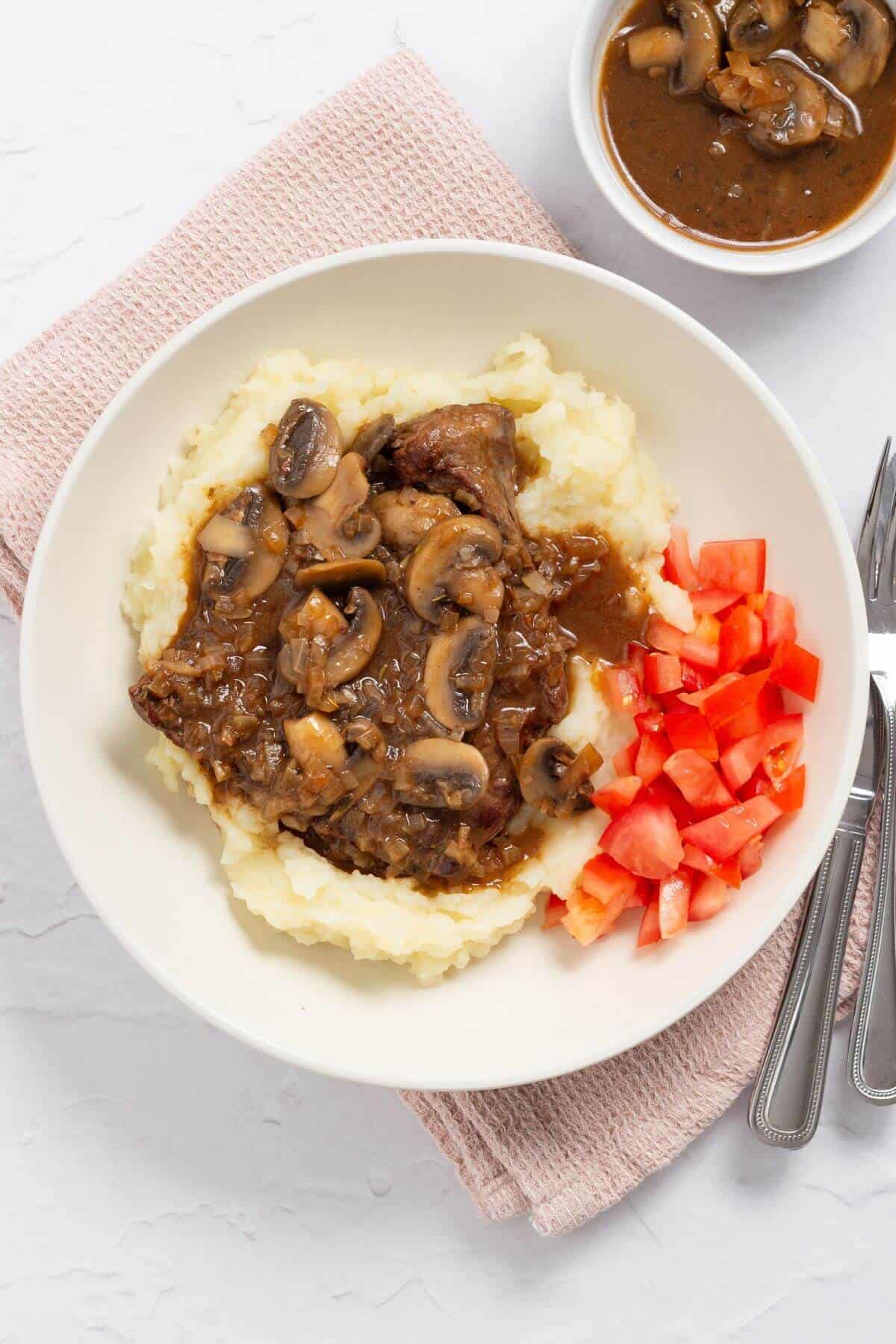 Mashed potatoes topped with beef, mushrooms, and gravy, served with diced tomatoes on the side; a bowl of gravy in the background.