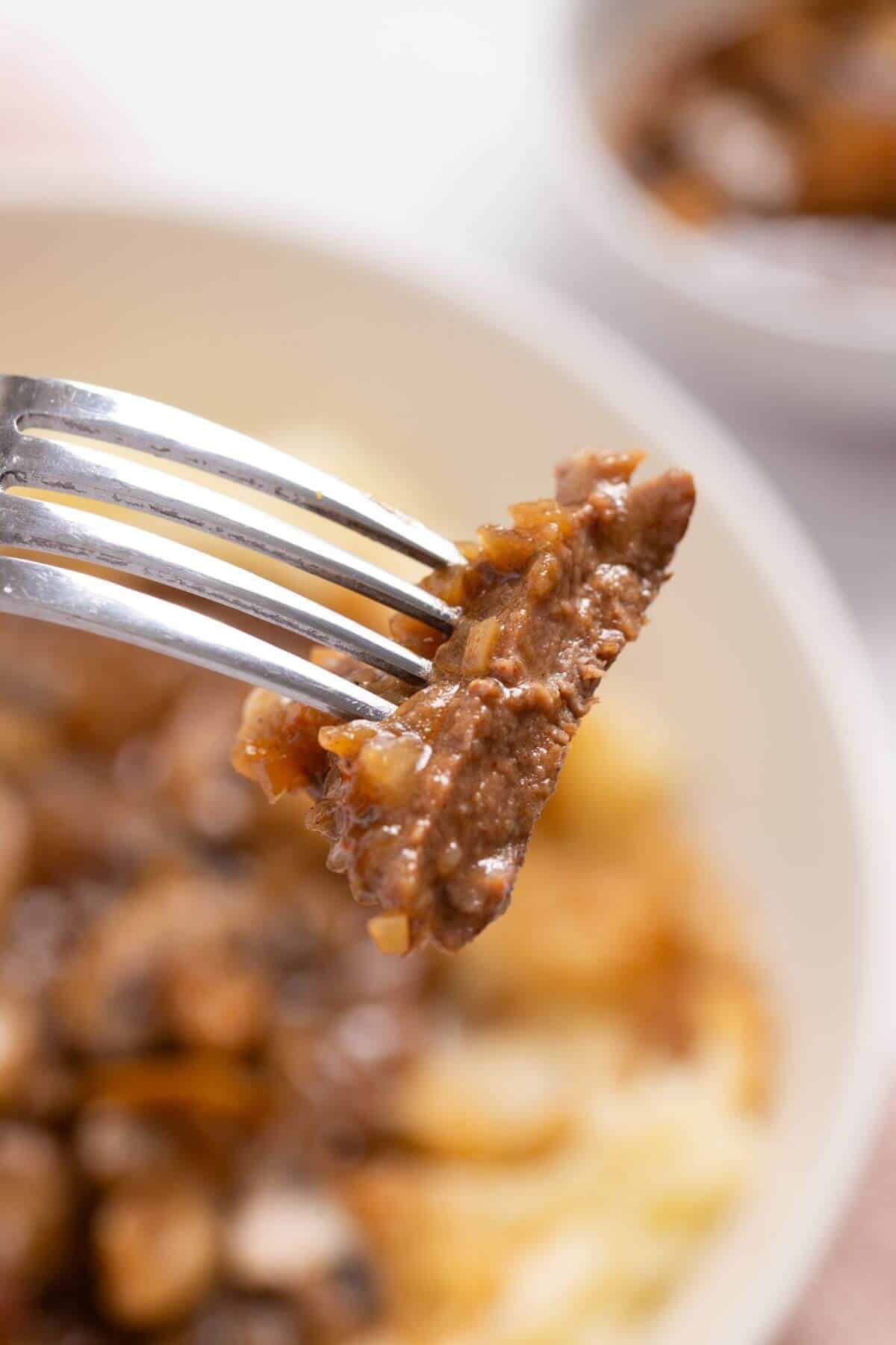 A fork holding a piece of beef with onions, with a bowl of beef and mushroom dish in the blurred background.