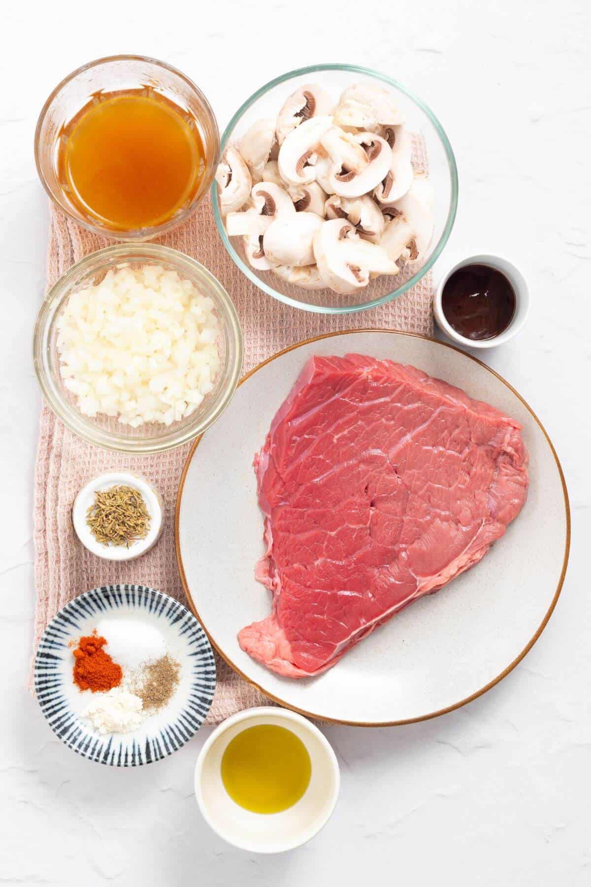 Plate with raw beef surrounded by bowls of sliced mushrooms, chopped onions, broth, seasoning, sauces, and oil on a textured surface.