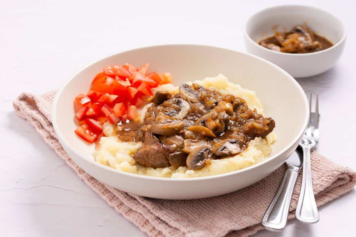 A plate with mashed potatoes topped with beef and mushroom gravy, diced tomatoes on the side, and a bowl of mushrooms in the background, placed on a beige cloth.