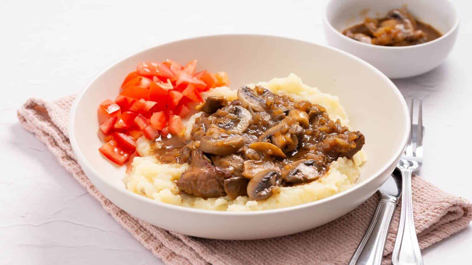 Plate with mashed potatoes topped with beef and mushroom gravy, alongside diced tomatoes. A bowl of extra gravy is in the background, with a fork and knife beside the plate.