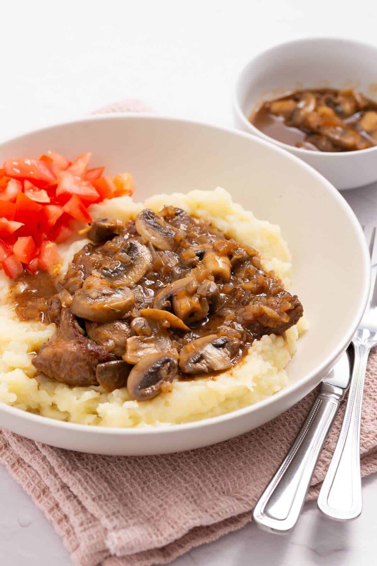 Plate with mashed potatoes topped with beef, mushrooms, and sauce. Diced tomatoes on the side. Bowl with extra sauce in the background. Fork and knife nearby.