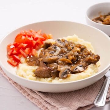 A bowl of mashed potatoes topped with beef and mushroom gravy, alongside diced tomatoes. A bowl of additional gravy is visible in the background.