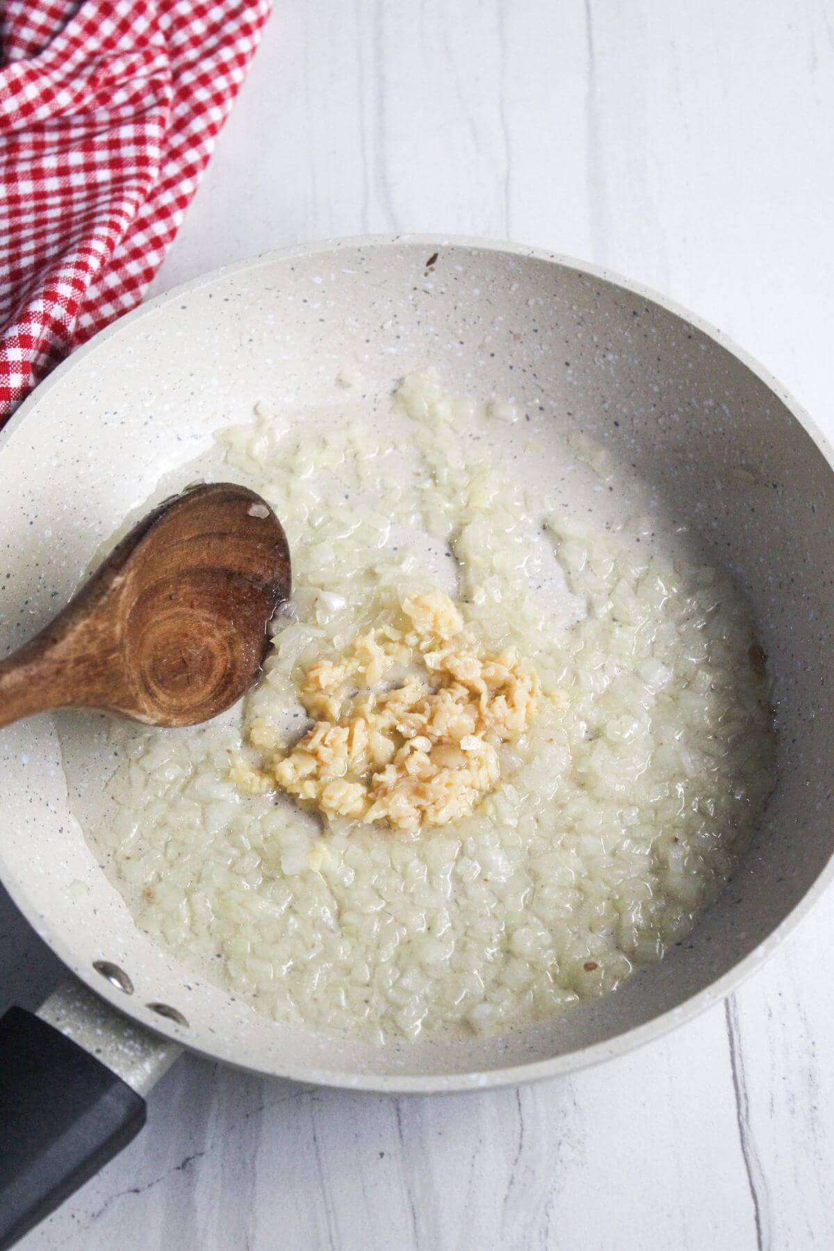 Chopped onions and minced garlic are sautéing in a white frying pan with a wooden spoon on a white countertop. A red and white checkered cloth is nearby.