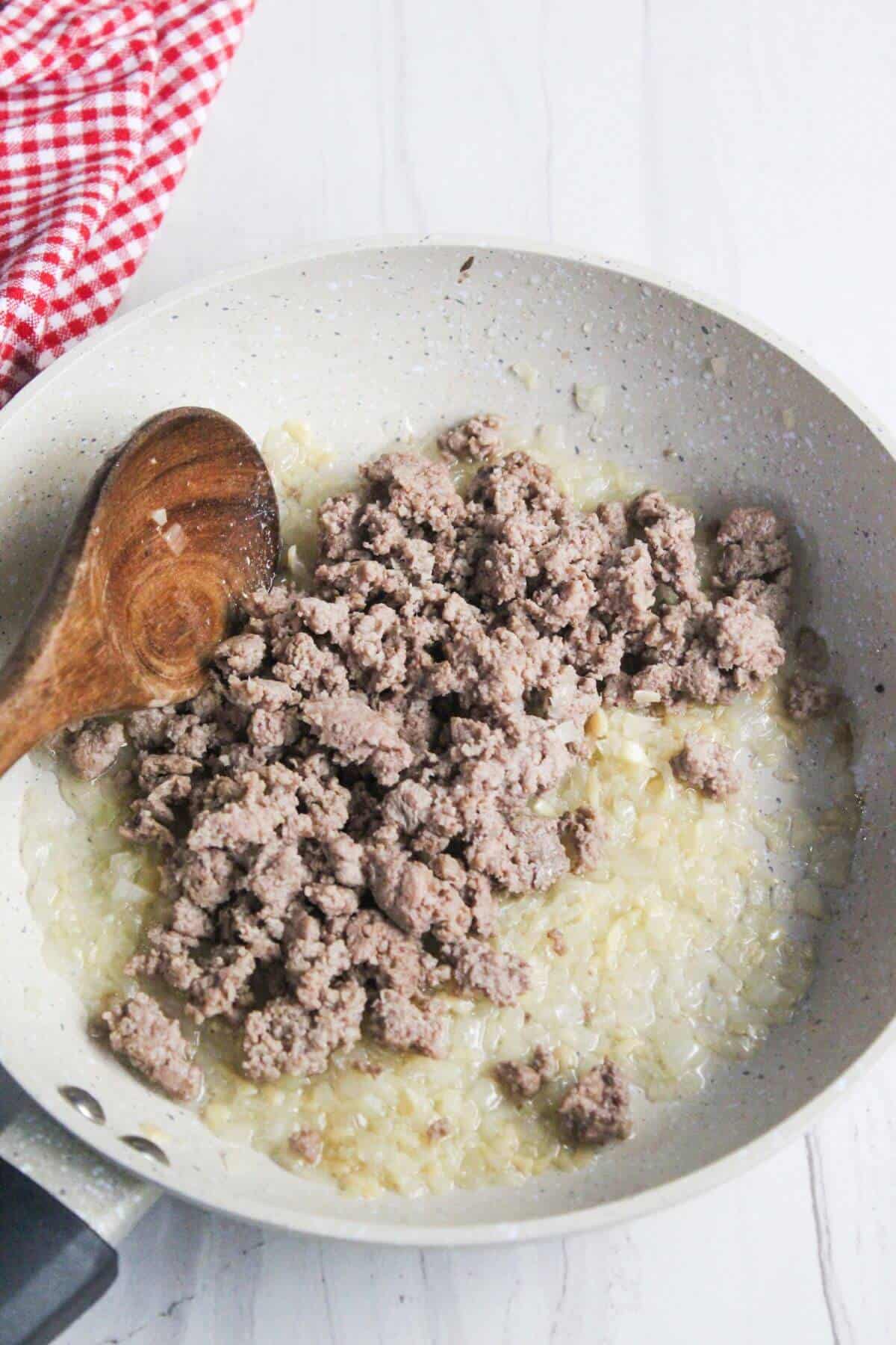 Ground beef and chopped onions cooking in a white skillet with a wooden spoon on the side.